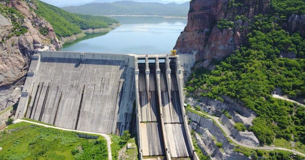 Represa en Sinaloa con compuertas abiertas y embalse al fondo, rodeado de colinas verdes.