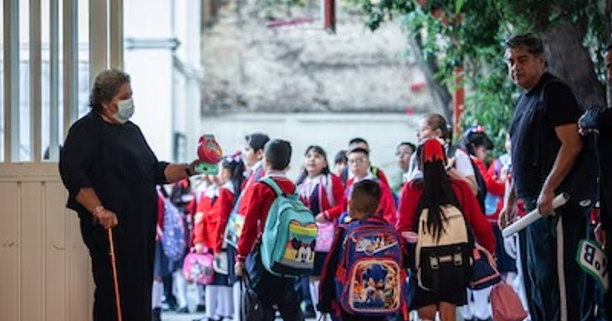 Niños en uniforme rojo con mochilas junto a una mujer con mascarilla, en un entorno escolar al aire libre.