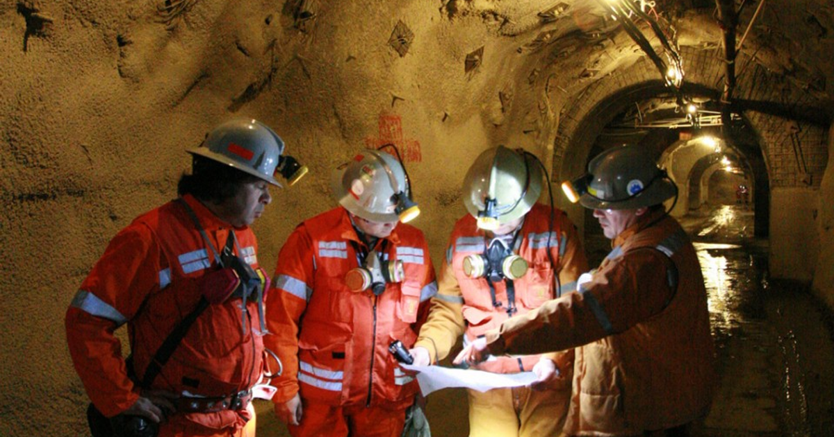 Trabajadores en túnel subterráneo con cascos y ropa naranja revisando planos, en el contexto del rescate en El Teniente.