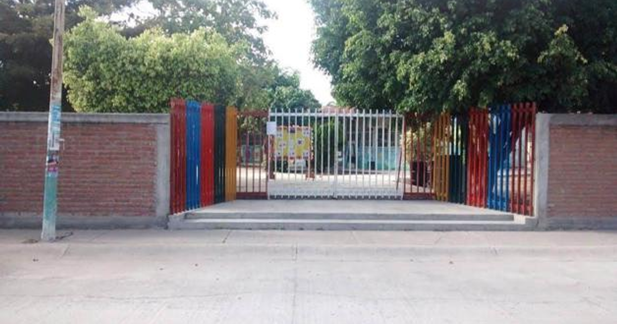 Verja de colores en escuela de Zapotillo, Ahome, con árboles y edificio al fondo.