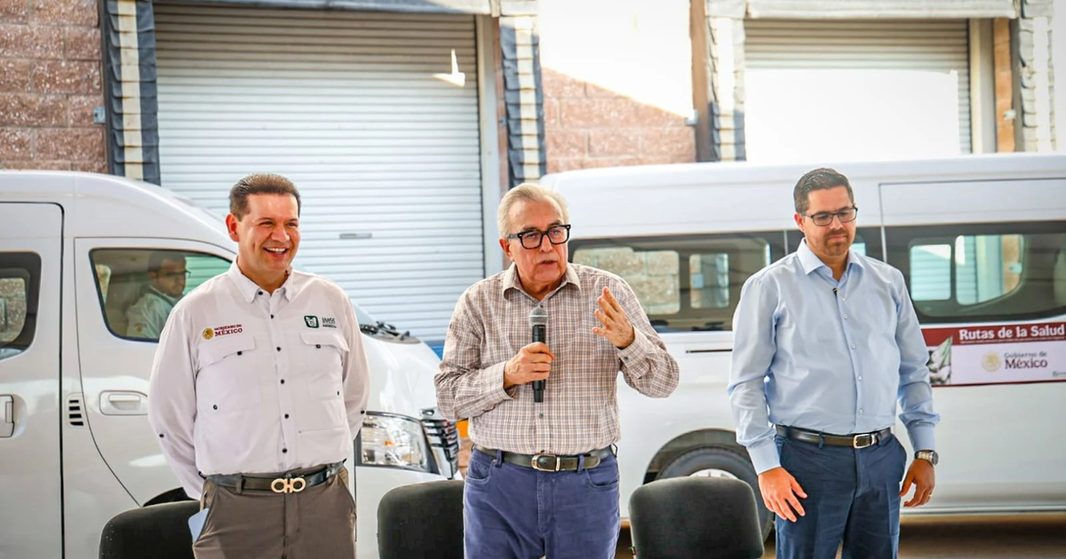 Tres personas en un evento del IMSS-Bienestar, una hablando al micrófono, con vehículos de salud al fondo.