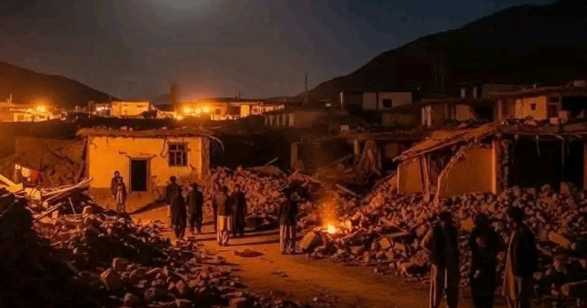 Paisaje nocturno devastado en Afganistán tras terremoto, con edificios en ruinas y personas reunidas alrededor de un fuego.