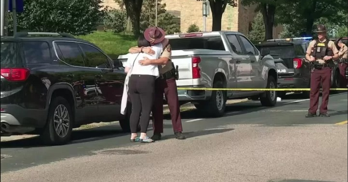 Oficial de policía abrazando a una mujer en escena de incidente con patrullas y cinta amarilla tras tiroteo en escuela de Minneapolis.