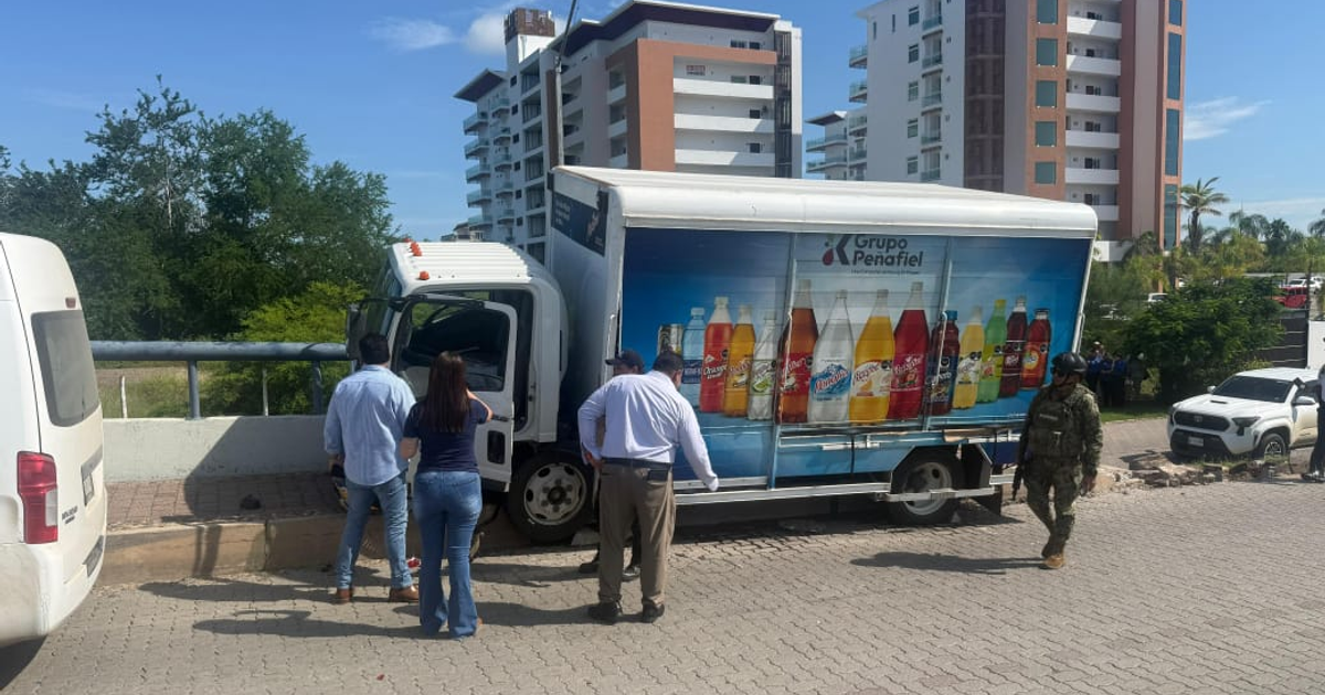 Camión de reparto de Grupo Peñafiel en escena de accidente en Mazatlán, rodeado de personas y un soldado, con un edificio y vegetación al fondo.