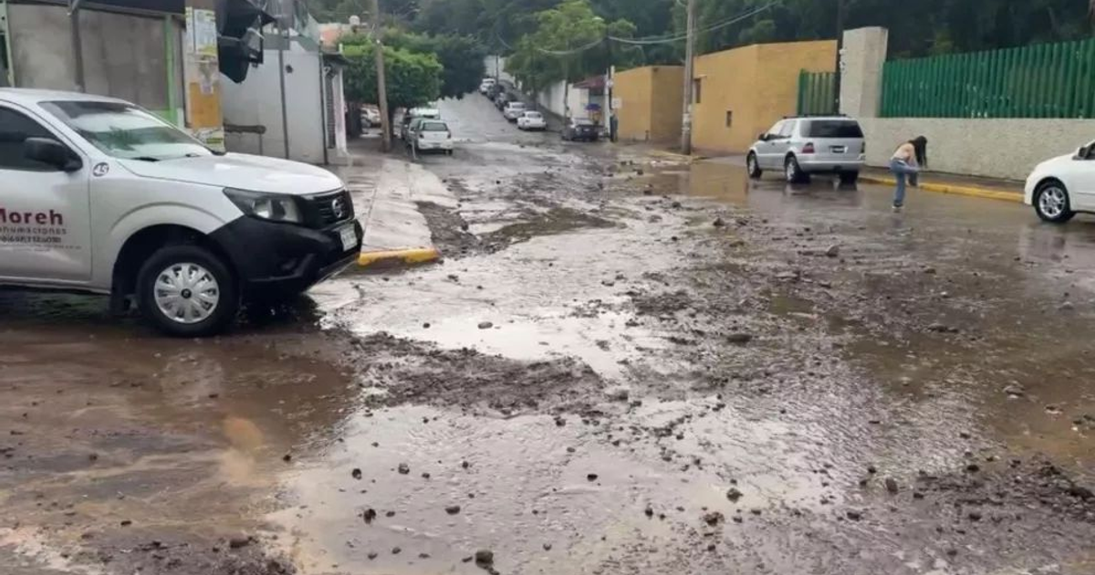Calle en Culiacán con charcos y lodo tras tormenta eléctrica, vehículos estacionados y una persona en el entorno.