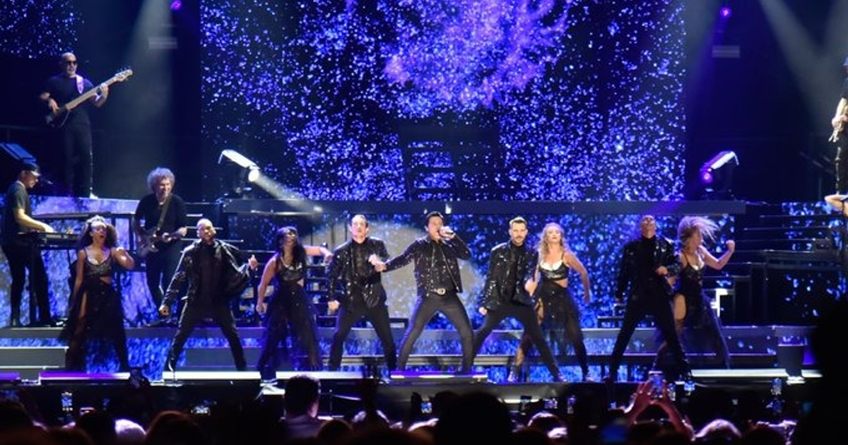 Chayanne actuando en el escenario de Chevron Park, rodeado de luces y efectos visuales, con una multitud de fans grabando el momento.