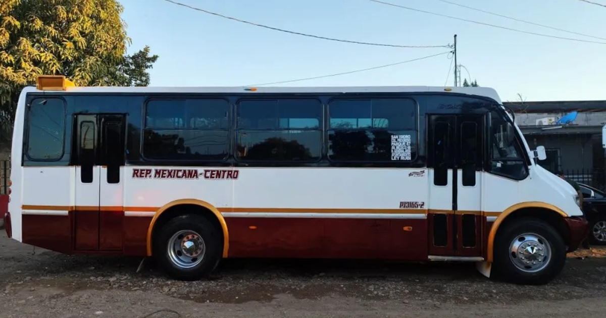 Autobús blanco y marrón de Culiacán estacionado en un área de tierra, con árboles y cables eléctricos al fondo.