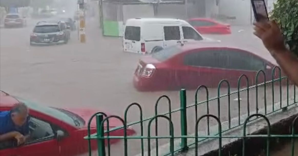 Vehículos sumergidos en una calle inundada bajo intensa lluvia, con un hombre tomando fotos.