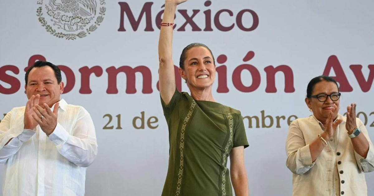 Claudia Sheinbaum en evento de Transformación Avanzada, levantando la mano y sonriendo, con dos personas aplaudiendo a su lado.