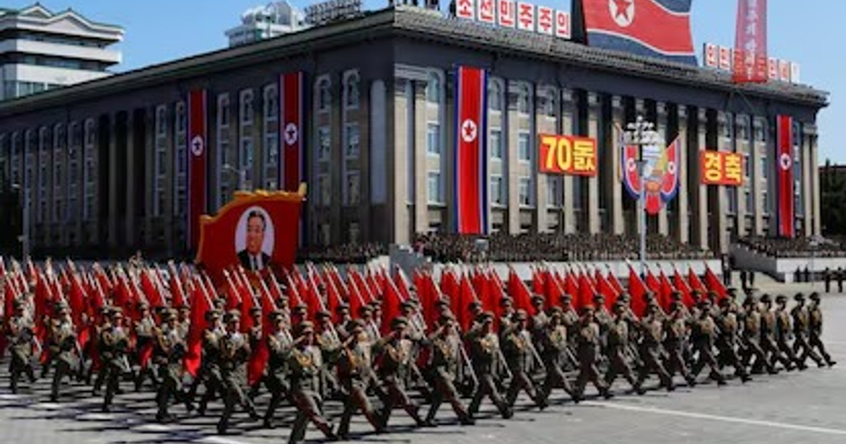 Soldados norcoreanos marchando con banderas rojas en un desfile militar, con un edificio decorado al fondo.
