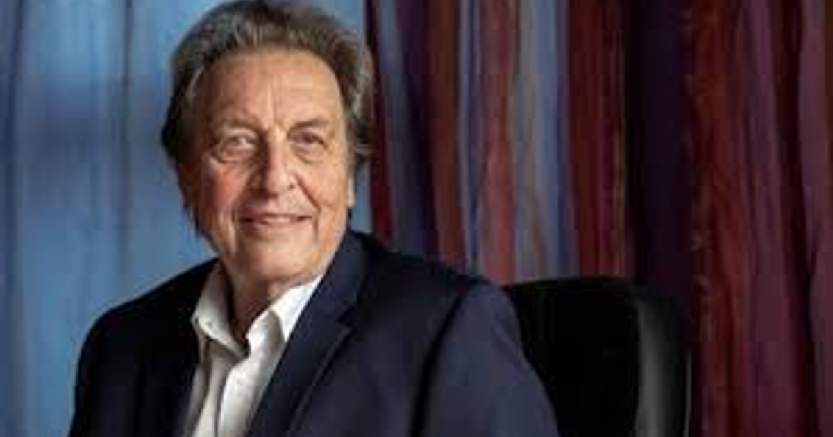 Hombre mayor con cabello canoso y traje oscuro sonriendo frente a cortinas de colores.