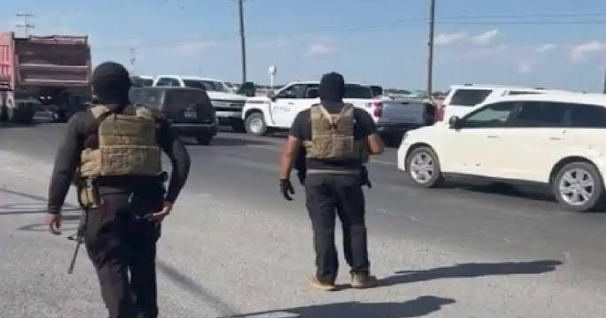 Personas con ropa táctica en carretera, rodeadas de vehículos, cielo despejado.