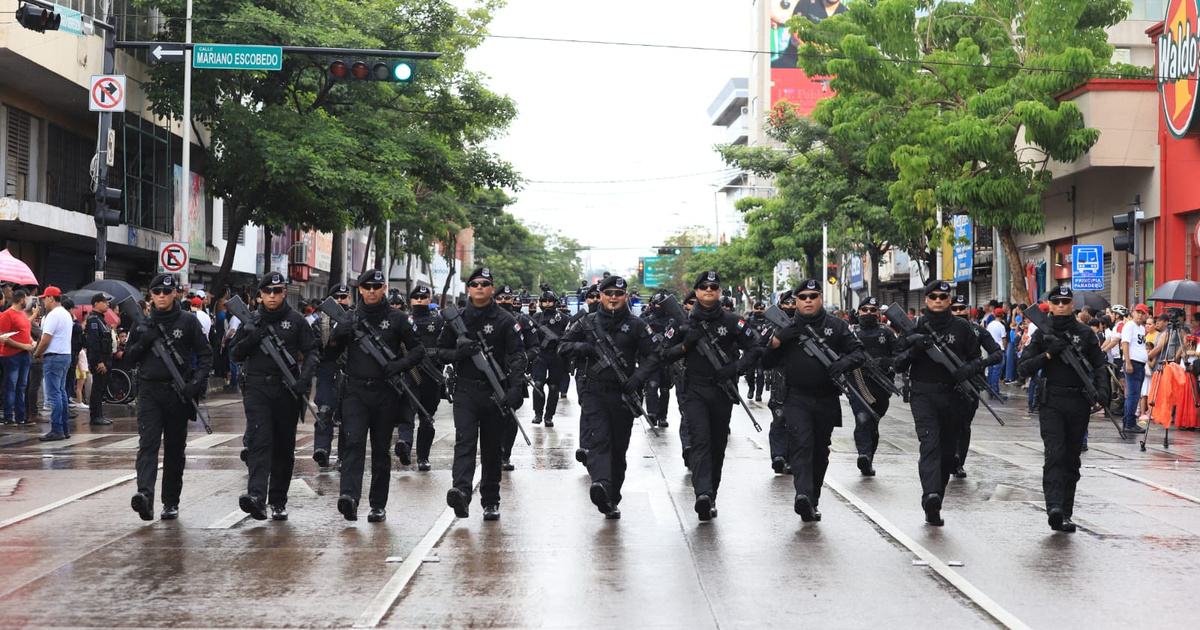 Oficiales del GOES marchando en Culiacán durante el desfile cívico-militar, con uniformes oscuros y armamento, bajo un clima lluvioso.
