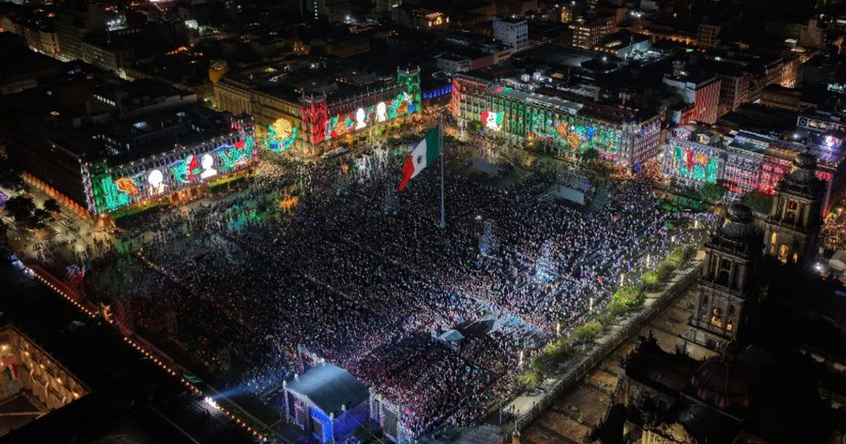 Multitud en el Zócalo de la Ciudad de México durante el Grito de Independencia, con luces festivas y una bandera de México.