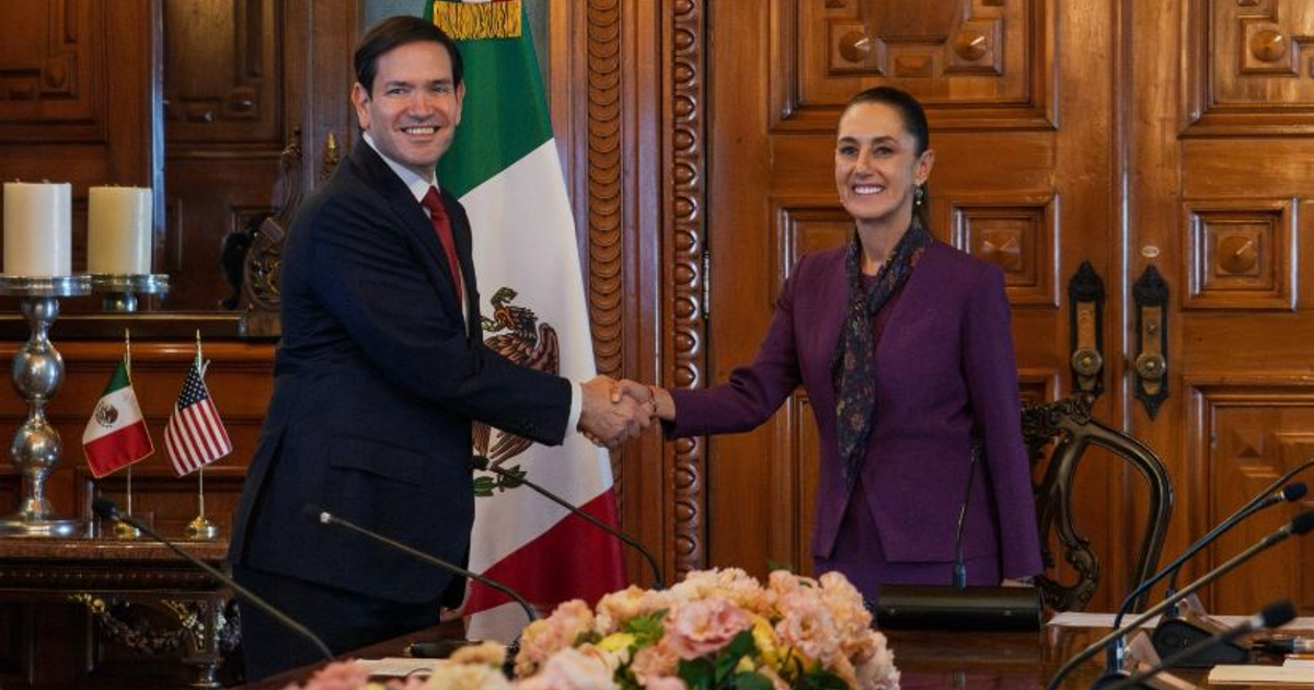 Dos personas estrechándose las manos frente a las banderas de México y Estados Unidos en un entorno formal.