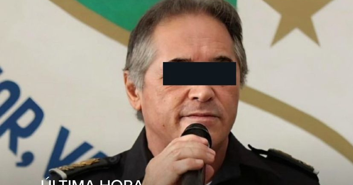 Hombre con micrófono en evento formal con bandera y emblema de fondo