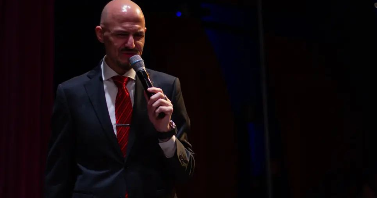 John Milton en el escenario con traje oscuro y corbata roja, sosteniendo un micrófono.