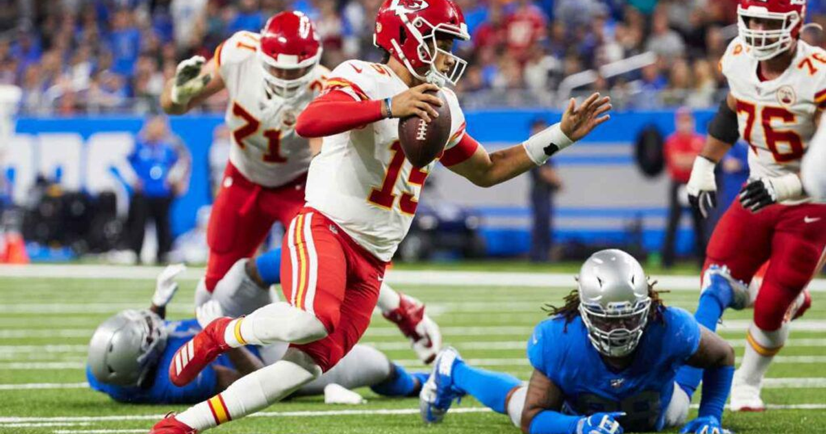 Jugador de fútbol americano lanzando el balón en un partido, con oponente en el suelo y público en las gradas.