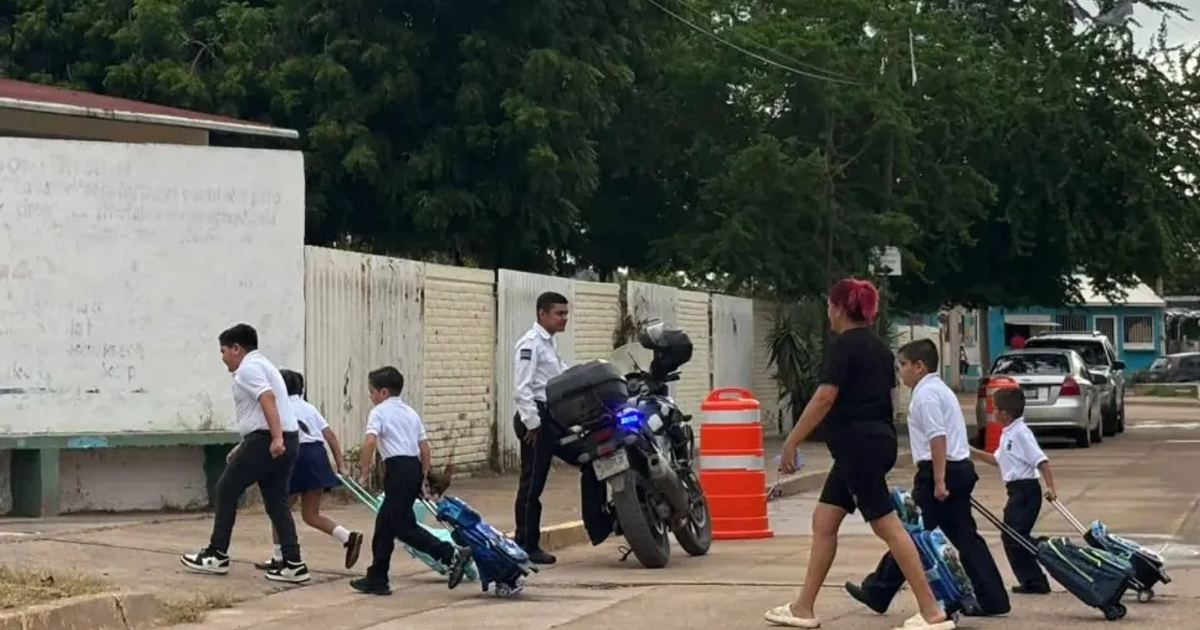 Niños con uniforme escolar caminando por la calle junto a un agente de seguridad en Mazatlán.