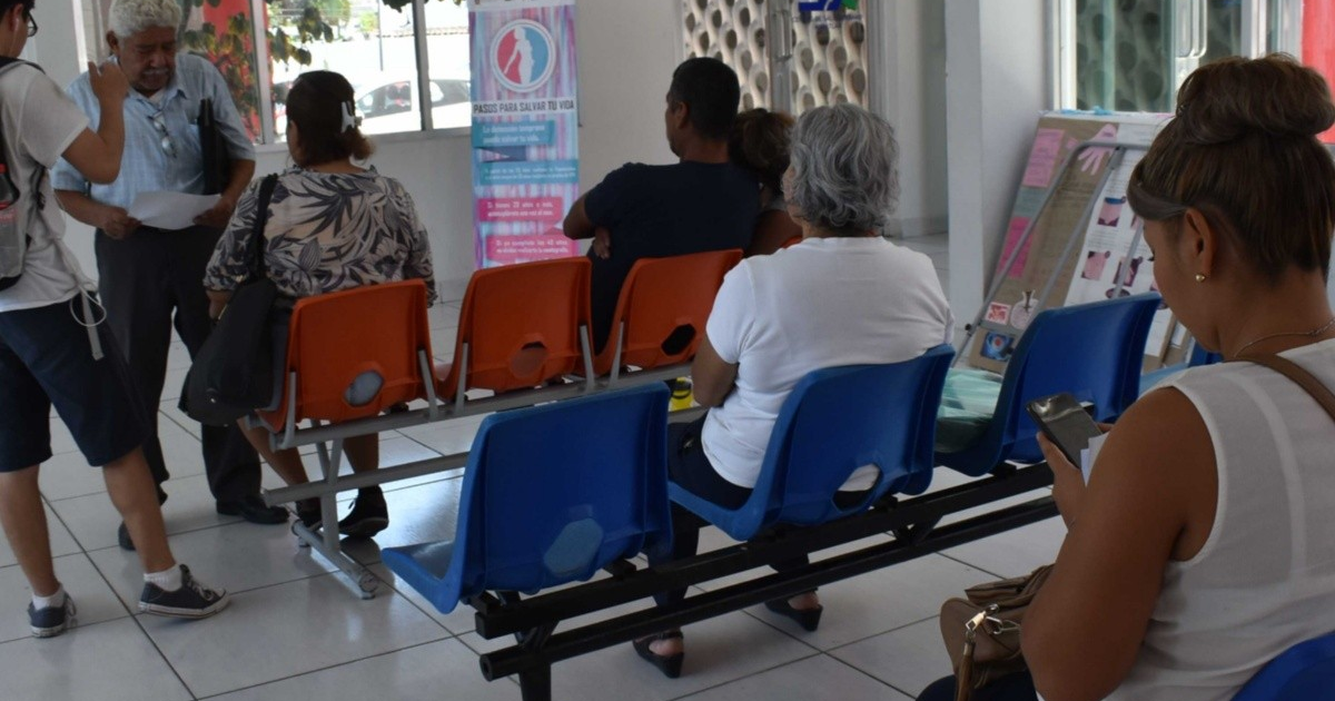 Personas en sala de espera durante evento de salud en Sinaloa, con bancos de colores y presentador de pie.