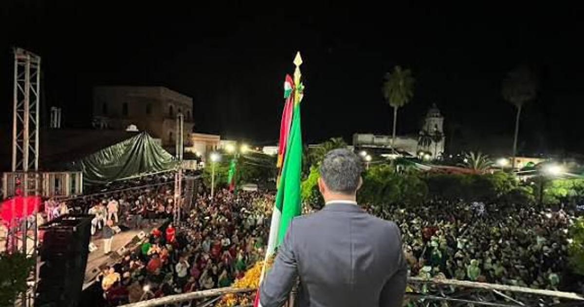 Orador con bandera mexicana frente a público en evento nocturno al aire libre