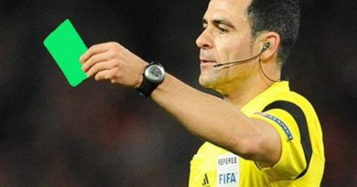 Árbitro de fútbol con tarjeta verde en mano durante un partido, con camiseta amarilla, auricular y reloj.