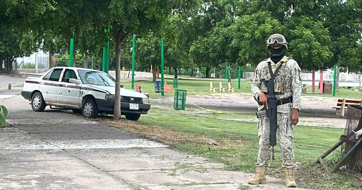 Soldado en Culiacán vigilando tras ataque a taxista, con auto dañado y árboles de fondo.