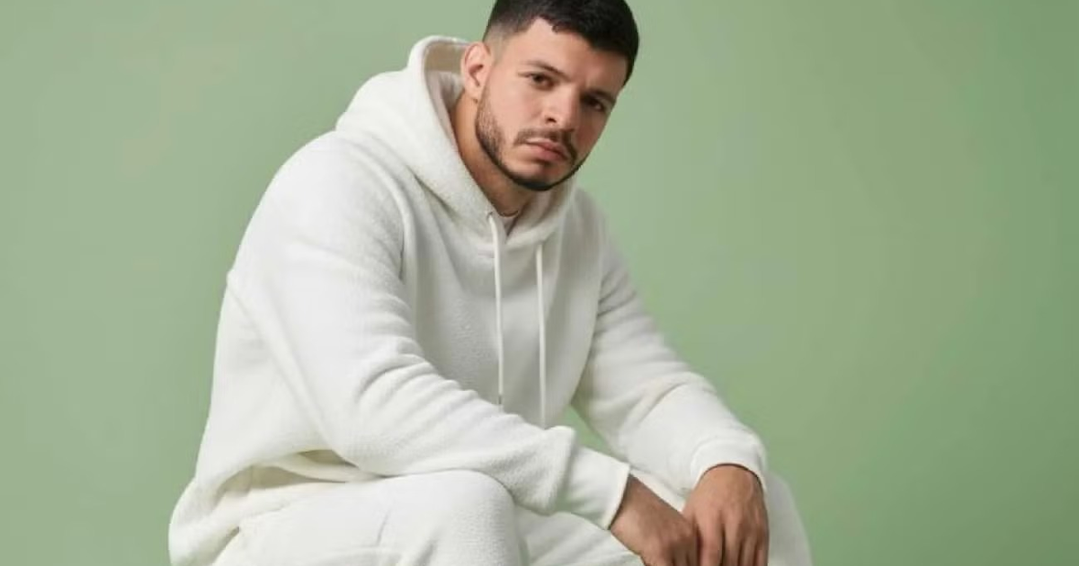 Tyron Paredes Gamboa, DJ venezolano desaparecido, sentado con conjunto blanco y fondo verde.