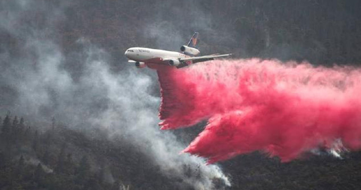 Avión lanzando retardante de fuego sobre un área con humo y vegetación.