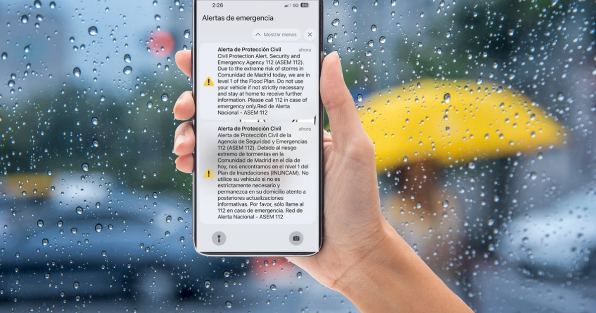 Teléfono móvil con alertas de emergencia climática en Madrid, fondo lluvioso y paraguas amarillo desenfocado.