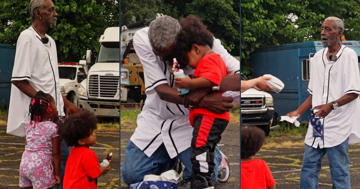 Hombre mayor abrazando a un niño con una niña cerca, en un entorno al aire libre con vehículos y árboles.