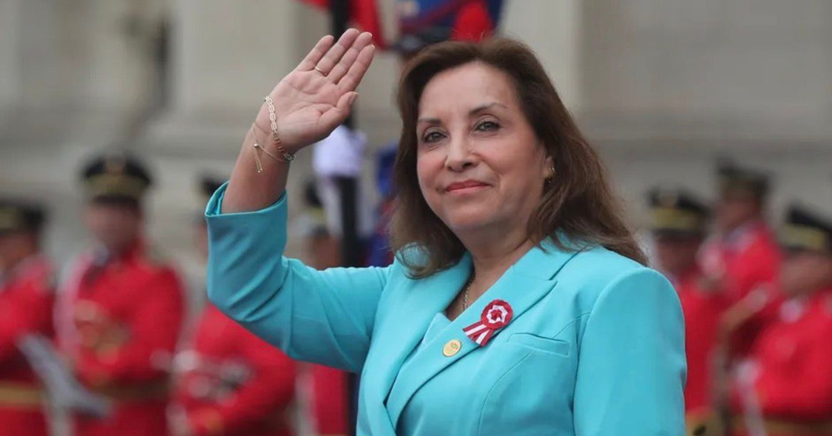 Dina Boluarte saludando en evento oficial con personas en uniforme rojo al fondo