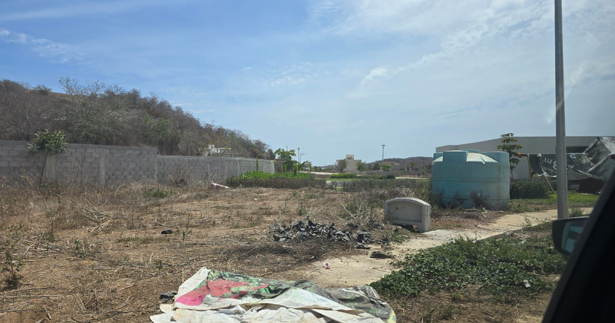 Terreno baldío en Veredas al Mar, Mazatlán, con muro de concreto, tanque azul y maleza.
