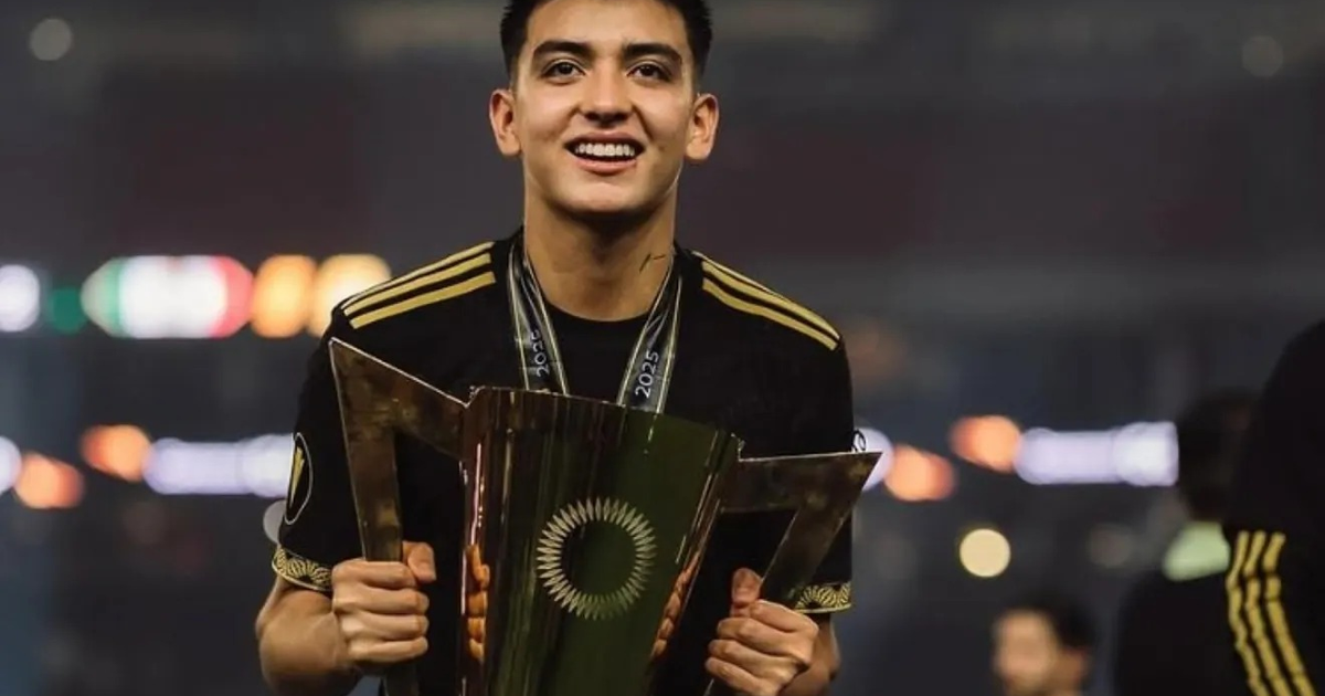Gilberto Mora sonríe sosteniendo un trofeo, vestido con camiseta negra y detalles dorados, en un entorno festivo.