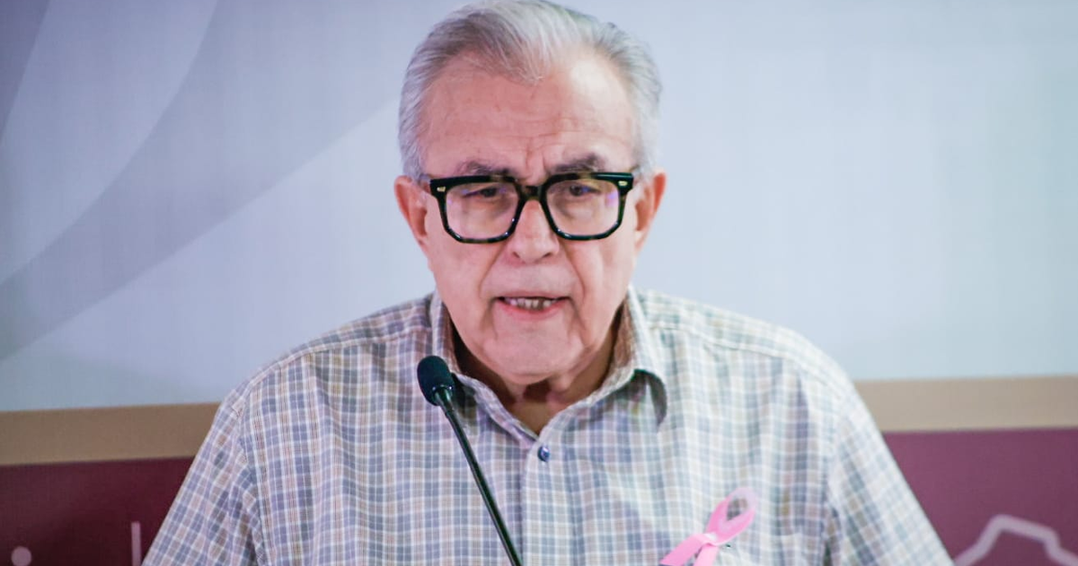 Hombre mayor con gafas y lazo rosa hablando en micrófono