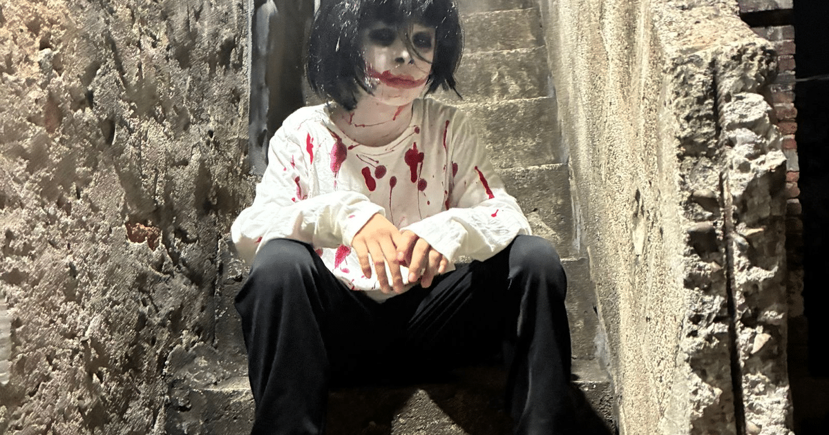 Persona disfrazada de Halloween con camiseta manchada de rojo y maquillaje de terror, sentada en una escalera.