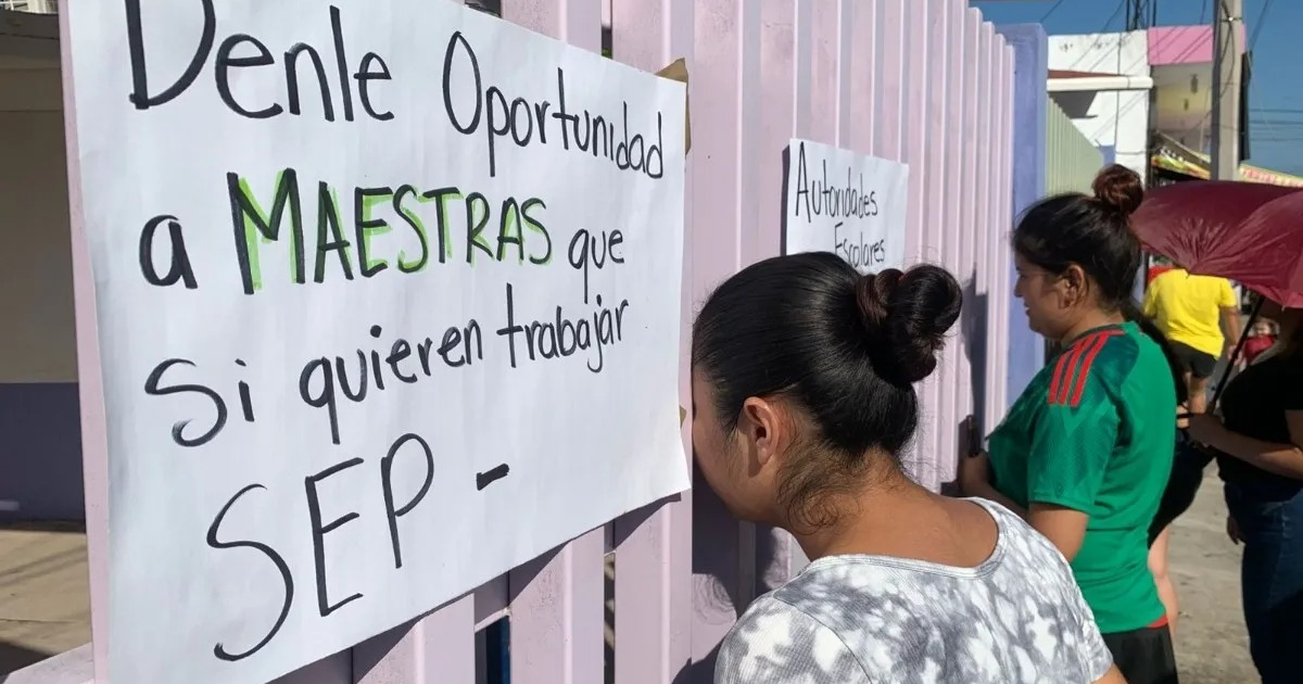Grupo de personas protestando frente a un edificio con carteles que exigen una maestra para el jardín de niños.