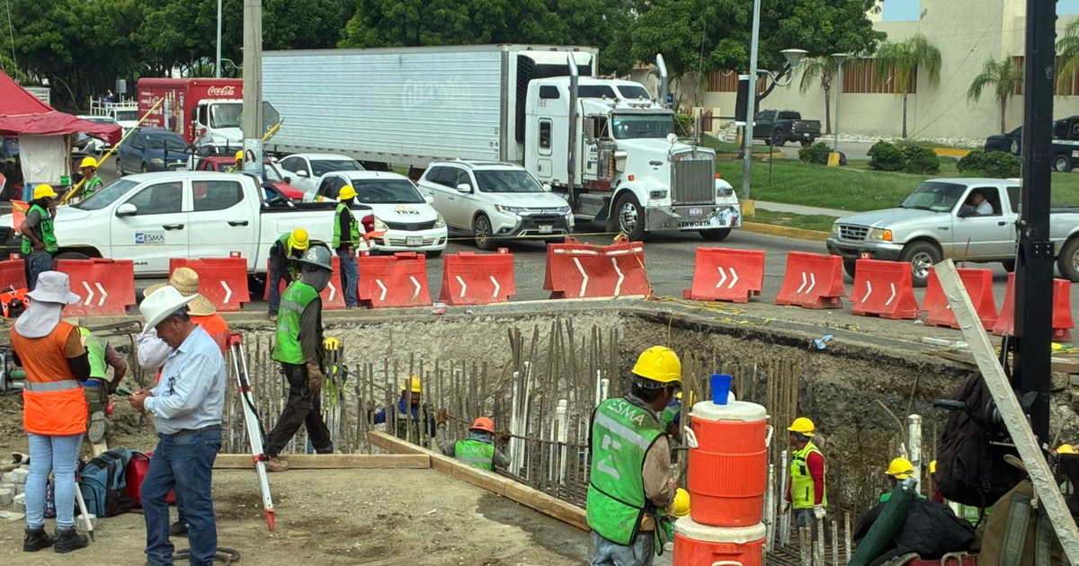 Trabajadores en la construcción del Puente Vehicular Norte en Mazatlán, con cascos y chalecos de seguridad, en un área delimitada por barreras de señalización.