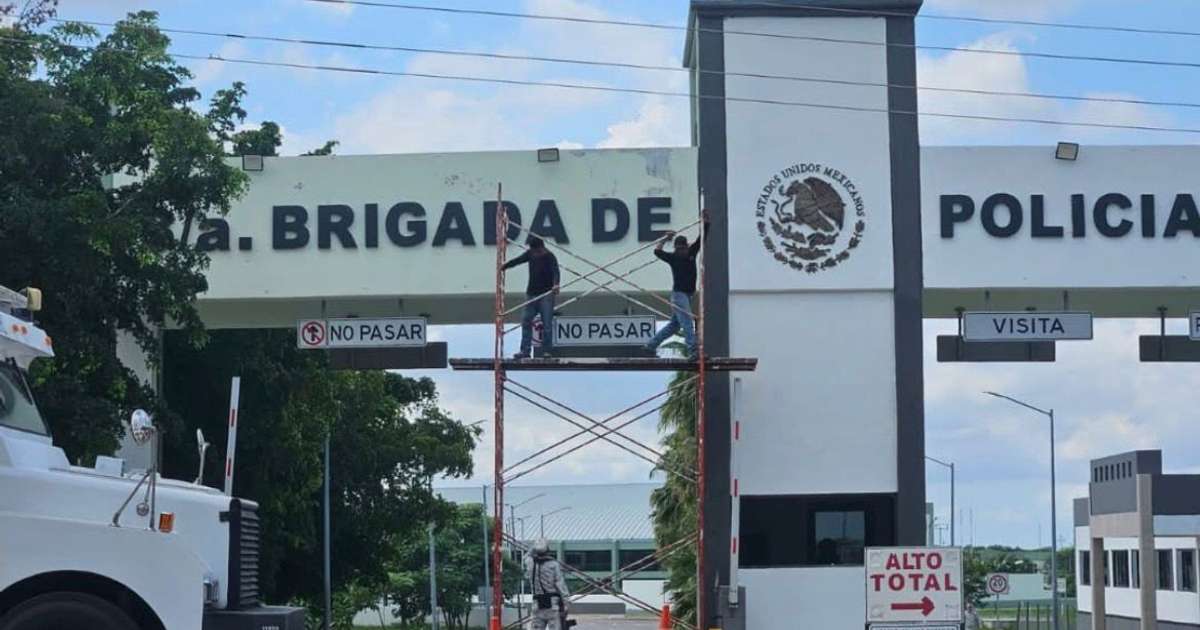 Trabajadores en andamio frente a portal de instalación policial en México, con letrero 'NO PASAR' y escudo de México visible.