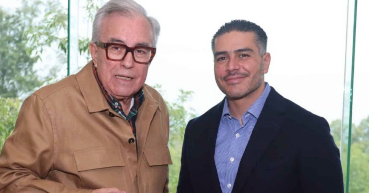 Dos personas sonrientes posando juntas en un interior con ventana y plantas de fondo.