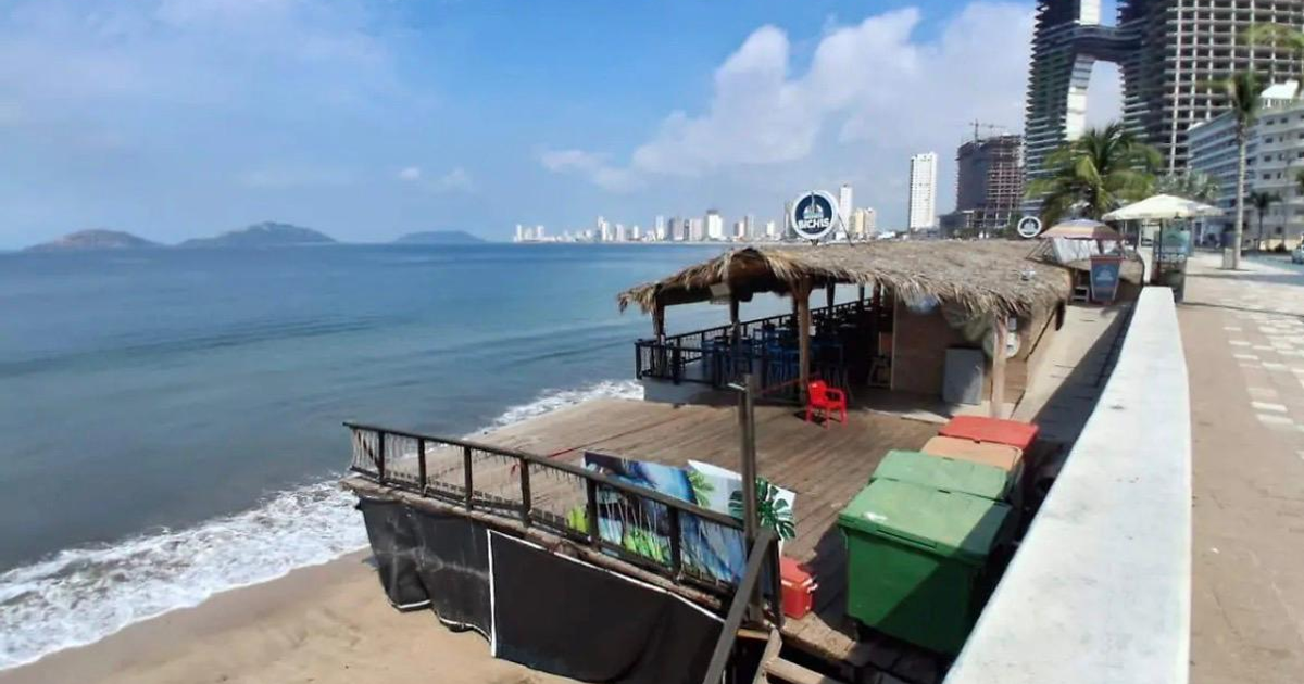 Playa en Mazatlán con palapa y muelle de madera, edificios al fondo.
