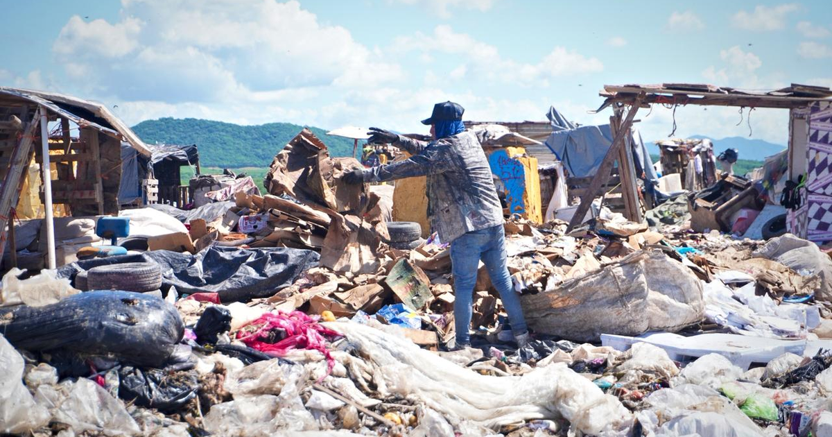Persona buscando entre basura en un paisaje desordenado con estructuras improvisadas al fondo.