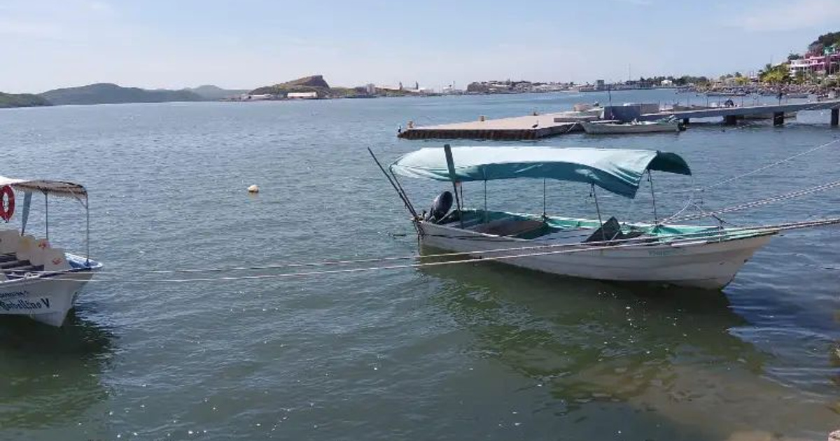 Bote con toldo azul anclado en el agua en Topolobampo, con muelle y montañas al fondo.