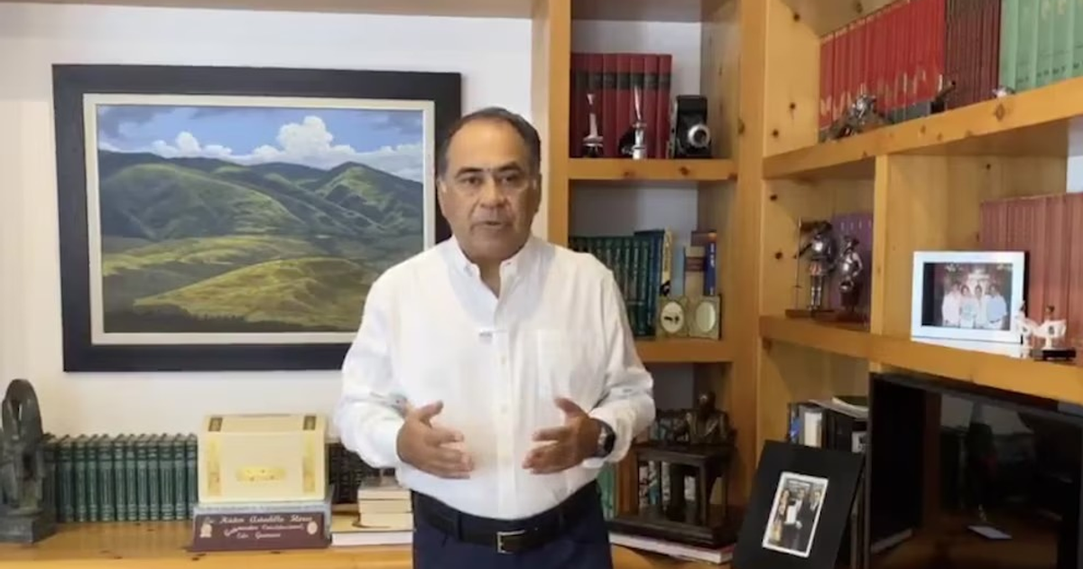 Persona de pie frente a estantería con libros verdes y objetos decorativos, explicando algo.