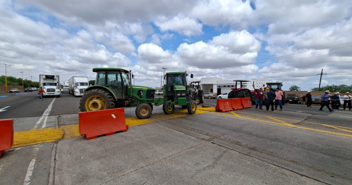 Tractores verdes bloqueando parcialmente una carretera en Guasave, Sinaloa, con barreras naranjas y personas interactuando.