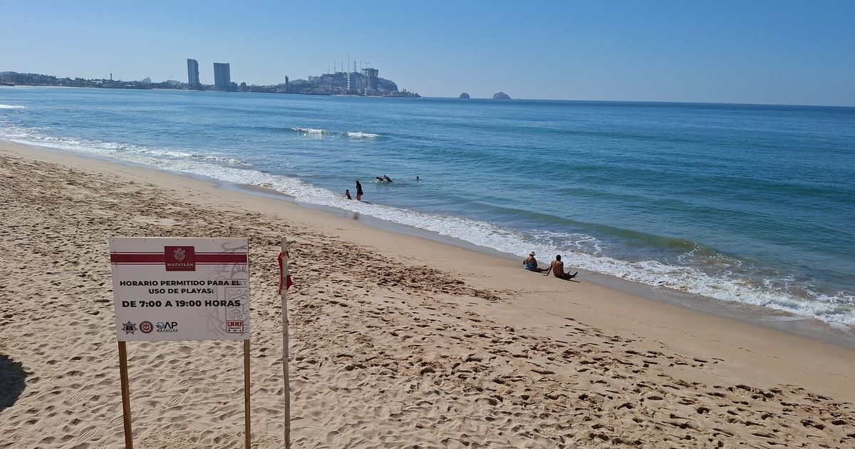 Playa de Mazatlán con letrero de horario y personas disfrutando del mar bajo un cielo despejado.