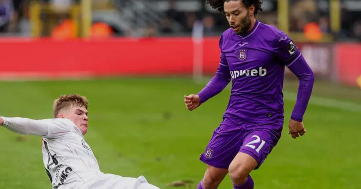 Jugador en blanco caído y jugador en púrpura corriendo en partido de fútbol con gradas de fondo
