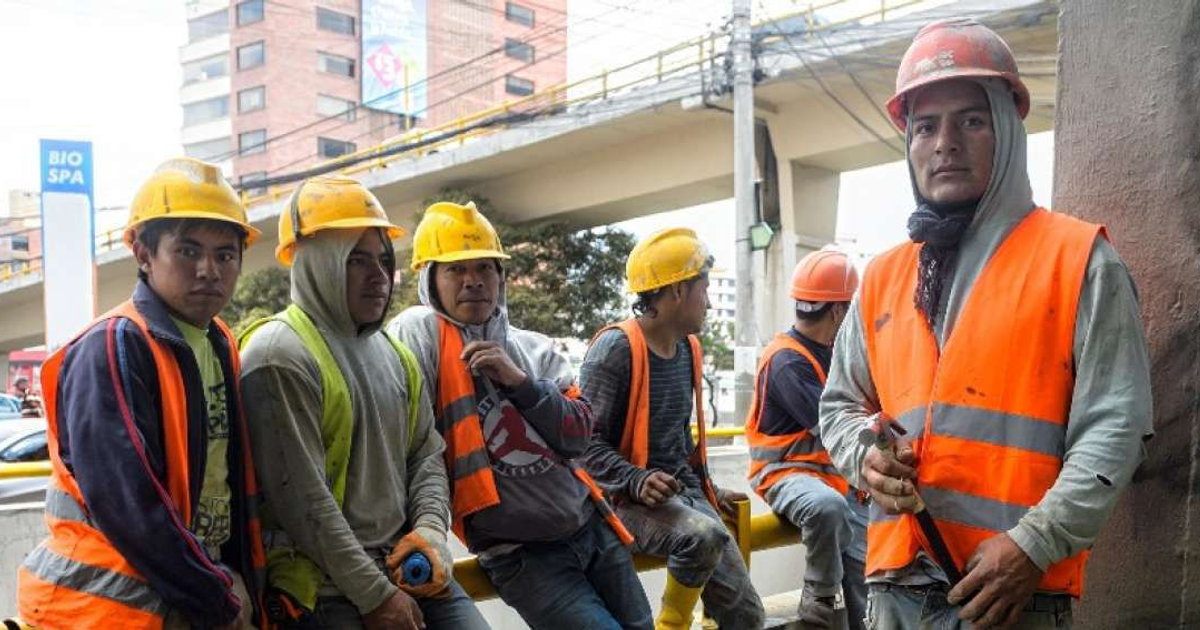 Grupo de trabajadores de la construcción con cascos amarillos y chalecos naranjas en un descanso, con un puente al fondo.