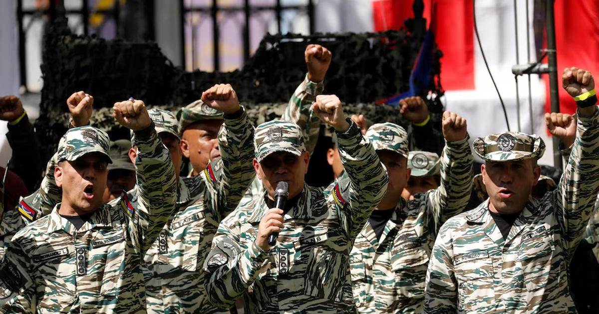 Hombres en uniformes militares de camuflaje levantando el puño en un evento oficial con banderas de fondo.