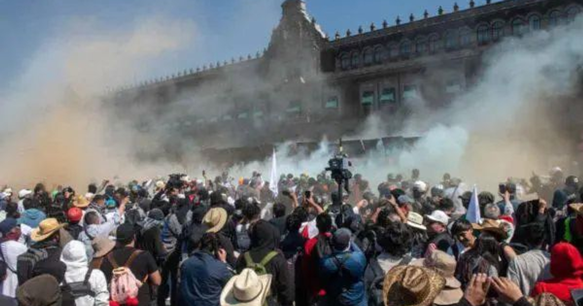 Multitud en el Zócalo durante manifestación con humo en el aire y edificio de fondo.